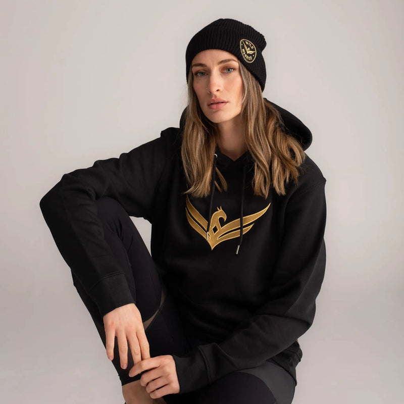 Woman wearing wild warrior pull over black hoodie with large gold logo embroidered on center and black beanie with badge logo embroidery